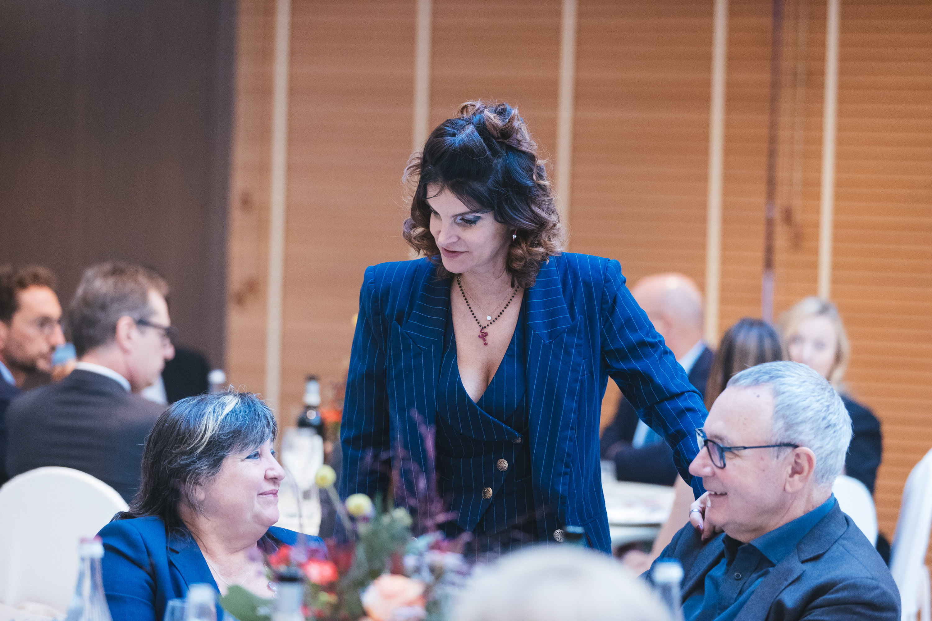 A white woman with blazer and waistcoat, dark hair, talking to people seated at a lunch table